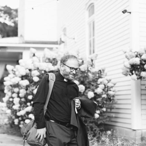 Pastor Brent, in his clergy collar, walks in front of a church.